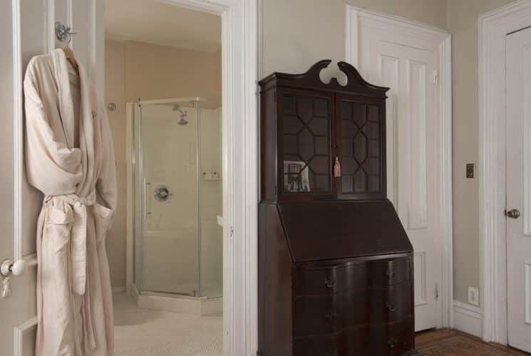 Bedroom with dark wood armoire and plush robe hanging on a doorway open to a bathroom with glass door shower